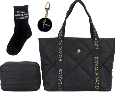 A black quilted Royal Academy of Dance tote bag with matching black accessories, including a black zippered pouch, a black sock with white text, and a round black keychain with a gold clasp, all displayed on a white background.